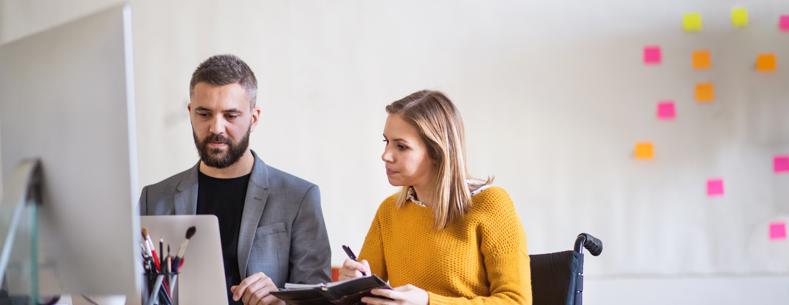 Dau berson yn eistedd wrth ddesg yn edrych ar gyfrifiadur. Mae un ohonyn nhw'n defnyddio cadair olwyn.