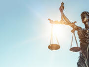 Cerflun efydd o Lady Justice yn dal clorian gytbwys yn ei llaw chwith a chleddyf yn ei llaw dde, gyda golau haul yn disgleirio trwy’r clorian yn erbyn awyr las glir.