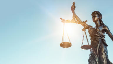 Cerflun efydd o Lady Justice yn dal clorian gytbwys yn ei llaw chwith a chleddyf yn ei llaw dde, gyda golau haul yn disgleirio trwy’r clorian yn erbyn awyr las glir.