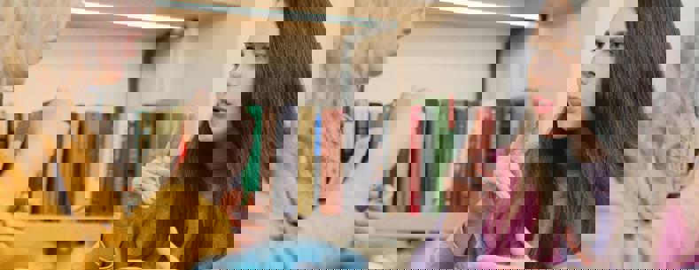 Dwy fenyw yn eistedd wrth fwrdd gyda phot o de, yn cael sgwrs drwy ddefnyddio Iaith Arwyddion Prydain.