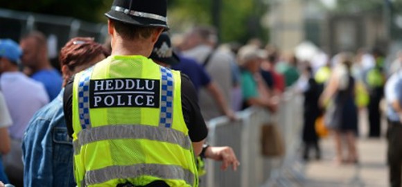 Swyddog heddlu mewn lifrai yn gwisgo fest felen welededd uchel, yn sefyll yn yr awyr agored, gan symboleiddio plismona rheng flaen ac amlygrwydd cyhoeddus. 