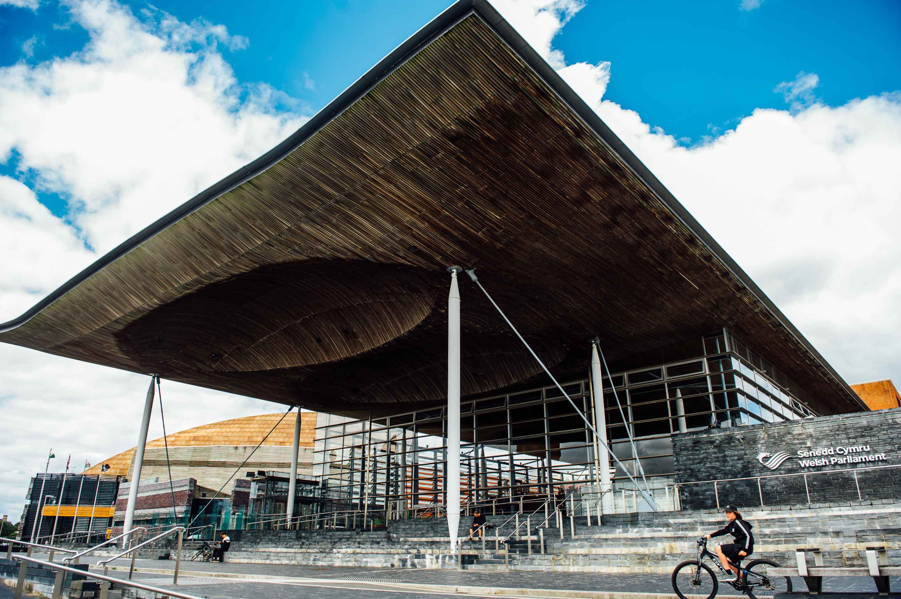 Delwedd o du allan adeilad y Senedd ym Mae Caerdydd, gydag awyr las a chymylau yn y cefndir. 