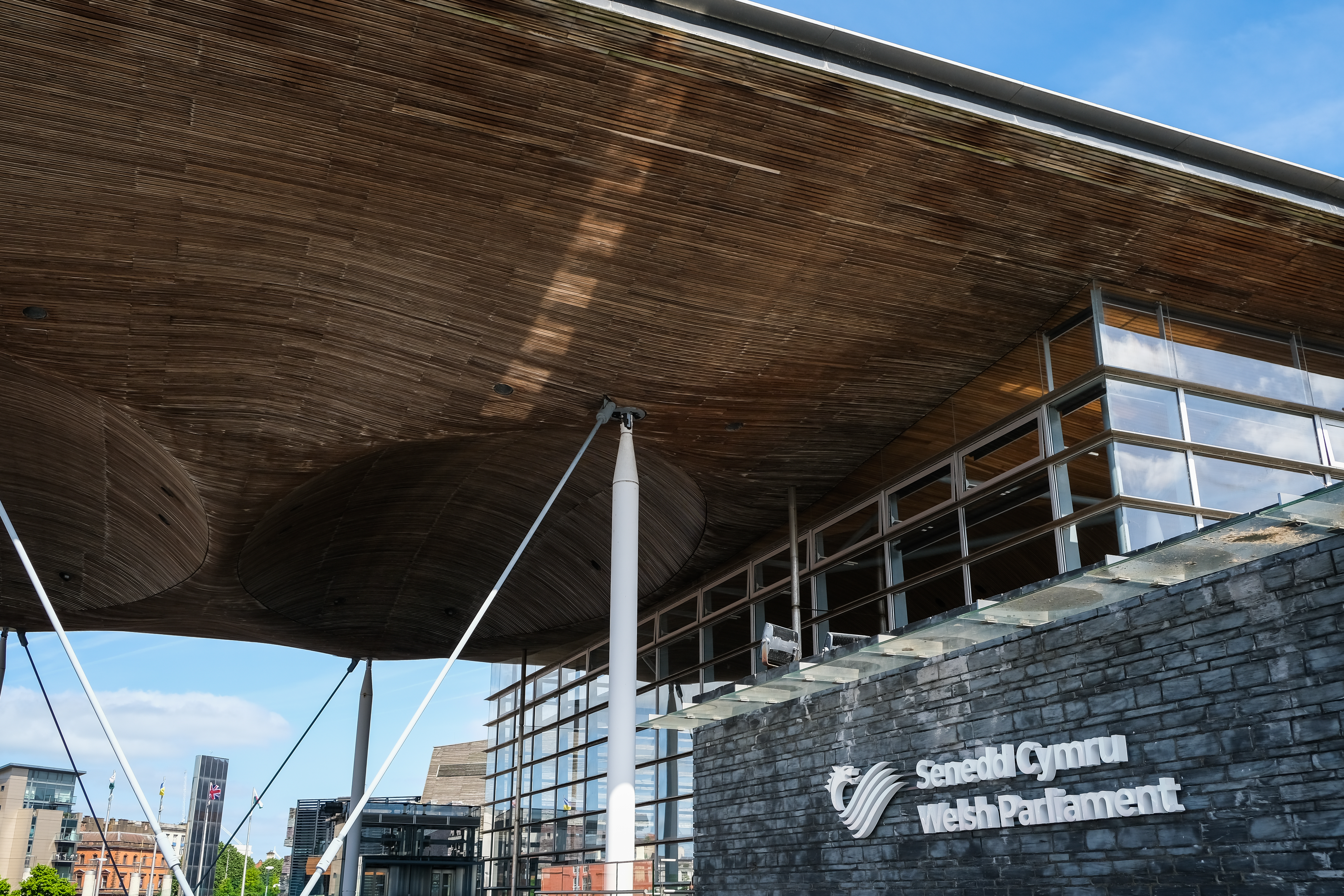 The exterior of the Senedd building in Cardiff Bay. The wooden roof takes up the top of the image and 'Senedd Cymru, Welsh Parliament' signage is visible on a wall in the bottom half of the image. Beams are connected to the roof and blue sky is visible in the top left corner.