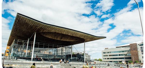 Ffotograff o du blaen adeilad y Senedd ym Mae Caerdydd. Mae'r adeilad wedi'i wneud o frics llwyd, ac mae rhai trawstiau gwyn i'w gweld. Mae arwydd sy'n dweud 'Senedd Cymru'. Yn y blaendir, mae cerddwyr a beicwyr yn mynd heibio. Mae rhai pobl yn eistedd ar y grisiau y tu allan i adeilad y Senedd.