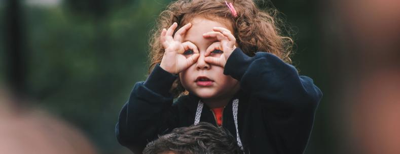 Girl sitting on an adult’s shoulders and using her hands to make binocular shape