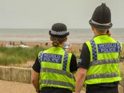 Dau swyddog heddlu mewn festiau ‘Heddlu Police’ yn edrych allan dros draeth gyda phobl yn y pellter.