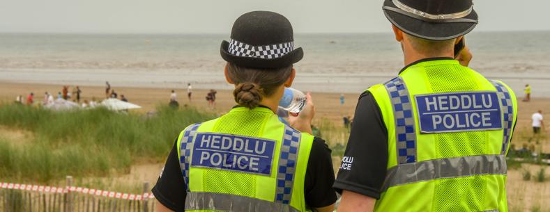 Dau swyddog heddlu mewn festiau ‘Heddlu Police’ yn edrych allan dros draeth gyda phobl yn y pellter.