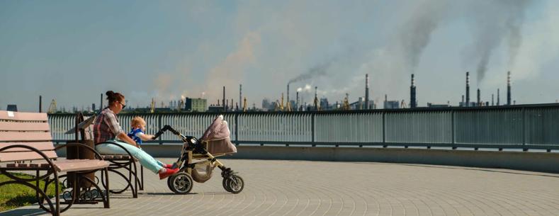 Menyw gyda phlentyn bach a phram yn eistedd ar fainc. Yn y pellter mae ffatri’n allyrru mygdarthau.