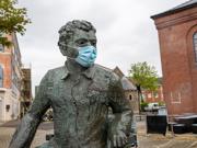 Cofeb efydd o Dylan Thomas yn eistedd mewn cwrt â blociau, yn gwisgo masg wyneb, gyda adeiladau brics a choed yn y cefndir.