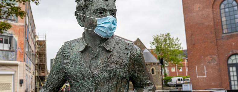 Cofeb efydd o Dylan Thomas yn eistedd mewn cwrt â blociau, yn gwisgo masg wyneb, gyda adeiladau brics a choed yn y cefndir.