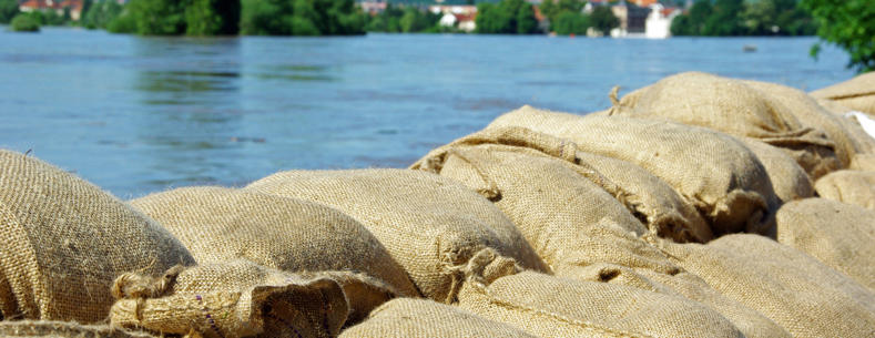 Ffotograff o fagiau tywod o flaen afon sydd wedi gorlifo.