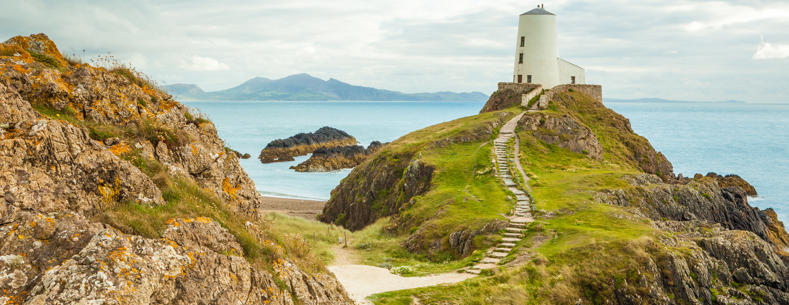 Ynys Llanddwyn, goleudy, gyda'r môr a'r mynyddoedd yn y cefndir.
