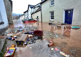 This is a picture of a street flooded by muddy water with floating rubbish and domestic items in the foreground. In the background are vehicles, most of which are submerged, one is  There is one floating car.