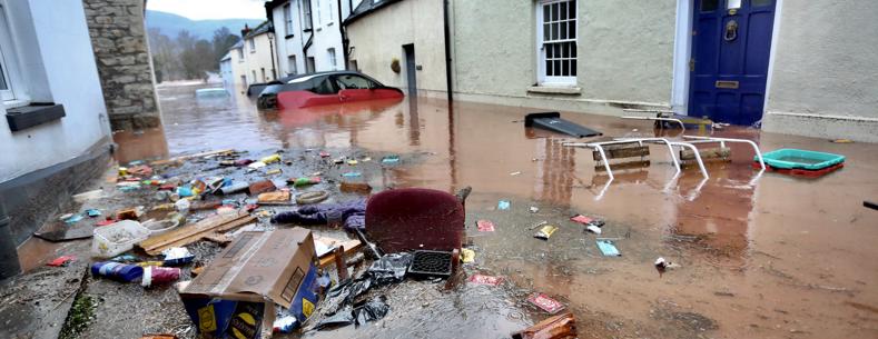 This is a picture of a street flooded by muddy water with floating rubbish and domestic items in the foreground. In the background are vehicles, most of which are submerged, one is  There is one floating car.